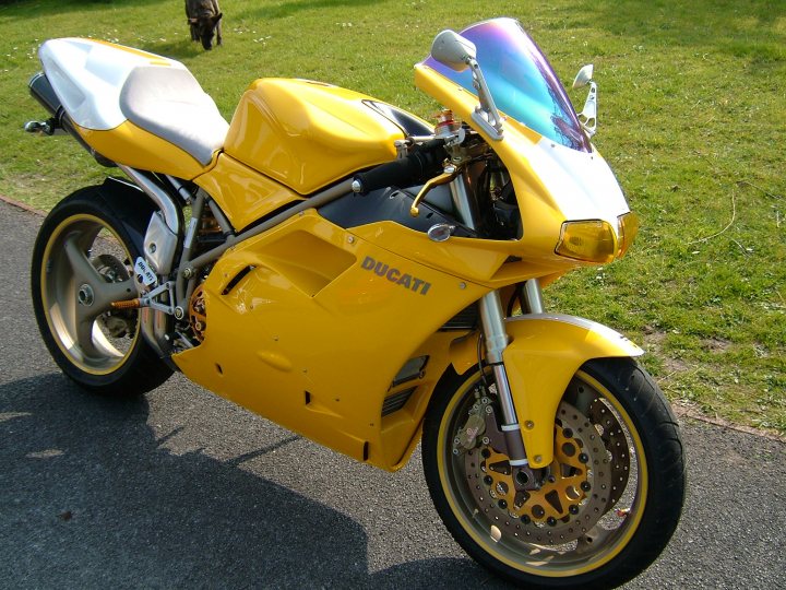 Any Bikers in here? - Page 1 - South West - PistonHeads - This image portrays a striking yellow DuCati motorcycle parked on a roadside against a backdrop of lush green grass. The motorcycle is positioned at a slight angle to the camera, showcasing its sleek design and reflective surface which is likely made up of various panels and components that gleam in the sunlight. 

The motorcycle is equipped with a fixed fairing and features familiar elements such as the round headlight, the elongated fuel tank, and the angular frame typical of the Ducati brand. The front wheel of the motorcycle is partially visible, where the spokes and the brake disc can be discerned. The motorcycle and the grass-covered field provide a vibrant contrast, with the motorcycle's bold color standing out against the tranquil and natural surroundings.