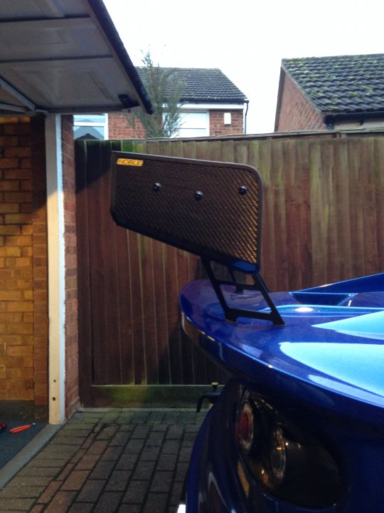 High Downforce Wing Risers - Page 1 - Noble - PistonHeads - The image captures a vibrant blue sports car parked on a brick driveway, adjacent to a residential house with a wooden fence. The car's black roll bar adds a touch of elegance and contrasts with the blue and red hues of the vehicle. Interestingly, a placard affixed to the roll bar suggests that the car may be participating in a time attack racing event, adding a sense of purpose and high-performance potential to the scene.