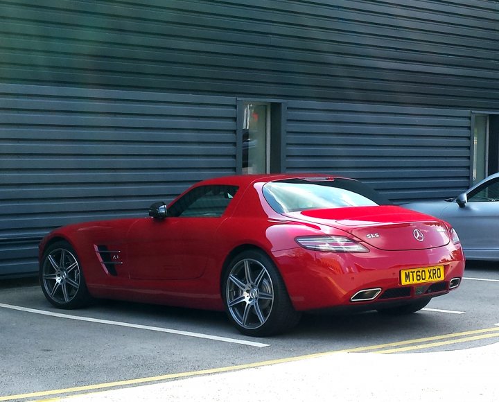 Supercars spotted, some rarities (vol 6) - Page 286 - General Gassing - PistonHeads - The image showcases a sleek red sports car parked in an open area with a building in the background. The car is positioned at an angle to the vehicle behind it, and its vibrant color contrasts sharply with the more subdued tones of the other car. The license plate is clearly visible, and the open area suggests a desolate, industrial setting. The reflection of the car indicates its shinny and well-kept exterior.