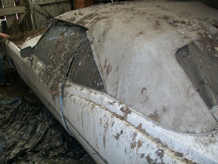 Shed Uncle Pistonheads - This image depicts a white classic car that appears to be relatively old, as evidenced by its worn and dirty exterior. The car's hood is up, revealing the interior structure and possibly a partial view of the engine. The body of the car shows signs of rust and grime, indicating it has been either neglected or stored in a poorly ventilated area. The car is parked on or under protective material like newspaper or discarded items, suggesting it is in a state of disrepair. The setting appears to be a garage or storage area with a wooden floor and metal walls, which is further reinforced by the presence of another similar car in the background.