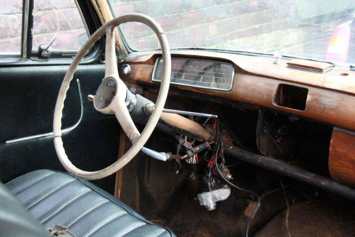 1950's Merc 220s Saved from the grave - rat rod restoration  - Page 1 - Mercedes - PistonHeads - The image is a photograph depicting an older model passenger vehicle's interior. The front driver's seat is filled with a variety of wires, which suggest that some form of repair, modification, or electrical work is being completed. The vehicle's dashboard is visible, with worn leather and wooden paneling providing a vintage aesthetic. The steering wheel is noticeable at the forefront of the image, with the driver's side door open, revealing glimpses of a brick building and what appears to be rain outside.