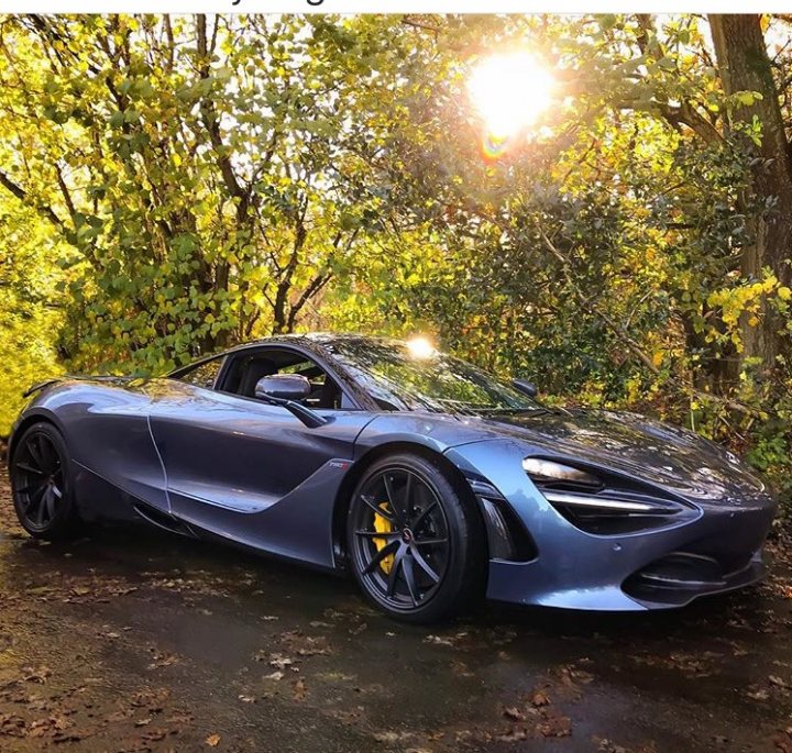 Just some photos of Mclarens - Page 1 - McLaren - PistonHeads - The image depicts a black McLaren sports car parked in a serene outdoor setting. The car's sleek design, with a low, aerodynamic body and large air intakes, is visible, and the vehicle features a large split front grille. The setting includes a lush, green environment, with trees and leaves decorating the background, creating a picture of a peaceful, wooded area. The sunlight filters through the trees and bounces off the shiny surface of the car, illuminating its features and adding a warm, natural backdrop to the scene. The overall image conveys a sense of luxury and tranquility.