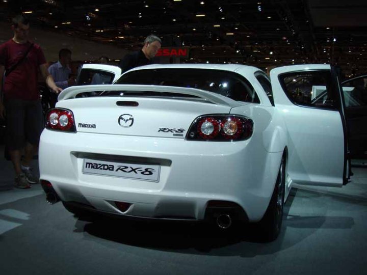 Pistonheads Revamp - In the heart of an indoor auto show, a Mazda RX8 car is the center of attention. The car is white, sleek, and elegantly parked facing the left side of the image. A man in the foreground is inspecting the design of this sports car, while another man stands attentively beside him, also studying the Mazda. This car exhibits a prominent red and black logo and multiple headlights, adding to its sporty charm. The setting is well-lit, emphasizing the car's colors and features, while the backdrop hints at more vehicles awaiting their turn in the spotlight.