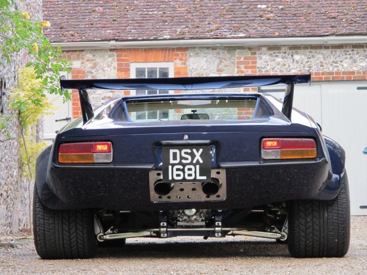 A car is parked in a parking lot - Pistonheads - The image shows a dark-colored sports car parked in a driveway next to a traditional British brick building. The car features a large rear wing and a number plate that says "D50X 168L." The car appears to be of a supercar caliber, with a sleek design that suggests speed and performance. The front lights of the car are illuminated, and a shield is visible on the hood. The car is positioned at an angle to the camera, displaying its striking features against the backdrop of the charming British house.