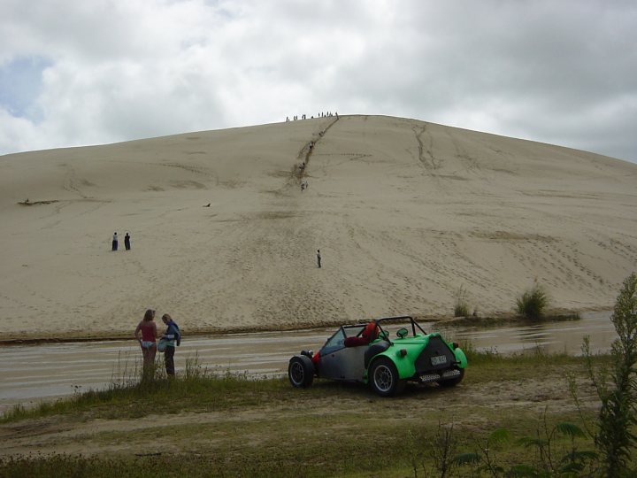 Beach Mile Pistonheads - The image features a green dune buggy parked near a sandy hill. In the foreground, a few individuals are standing and observing the vehicle with a mix of interest and surprise. Beyond the buggy and the individuals, a large sandy hill rises, and at its summit, several more people can be seen standing and walking. The scene appears to be set on a cloudy day, as evidenced by the grayish sky.