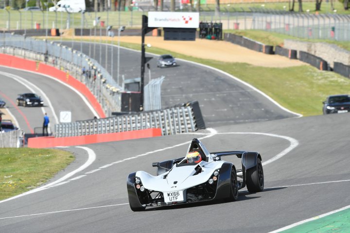 A motorcycle is parked on the side of the road - The image is a photograph taken during the daytime, showing a race car driver competing on a Formula One track. A sports car with open-wheel architecture and a prominent rear wing is prominently featured in the foreground, speeding down the track with visible motion blur. The car has a partial body wrap that appears to be a sponsorship deal with various logos. In the background, there are fencing barriers and other racing cars, suggesting an active race environment. The track itself is wide, well-paved, and appears to have multiple lanes for racing, with clear markings. There are no people visible in the image, focusing on the car and the racing environment.