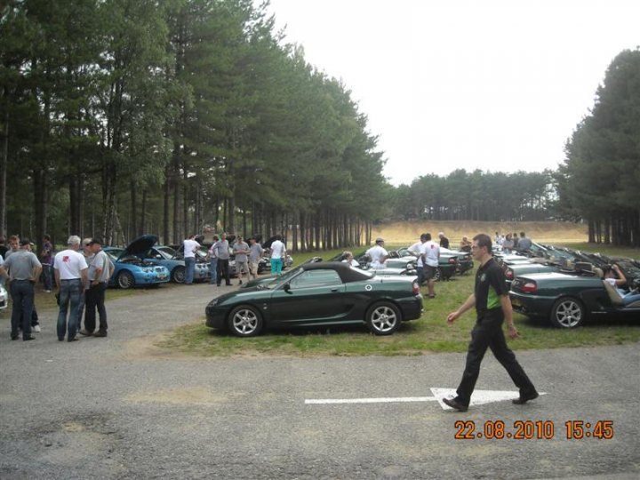 Pistonheads - The image displays an outdoor scene with a parking lot in the foreground where several people are standing near multiple cars. The vehicles vary in color and model, suggesting a casual gathering or event. The group of individuals appears to be either discussing or simply standing near their cars, contributing to a communal atmosphere. The area is surrounded by trees and is bordered by a grassy terrain, indicating a park or outdoor recreational area. The weather seems pleasant, and there are no visible clouds indicating a sunny day.