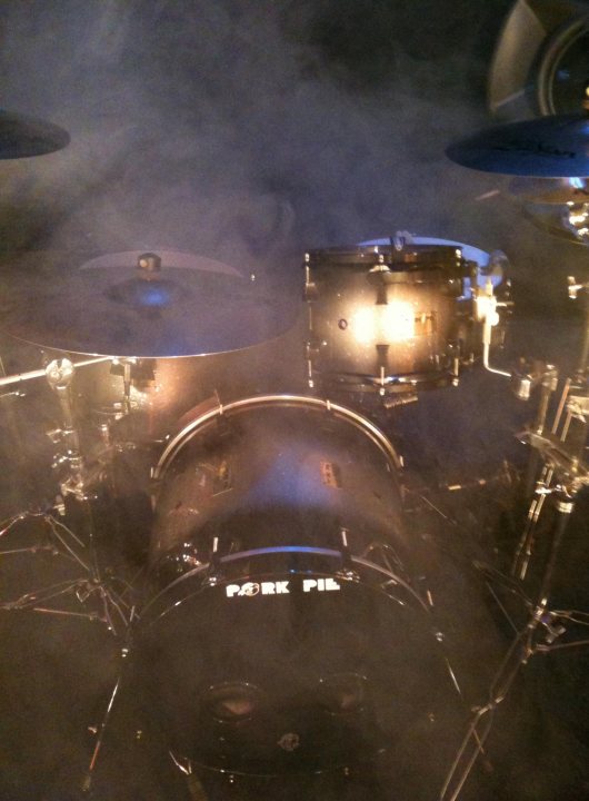 Pistonheads Instruments - The image captures the interior of a large drum kit, viewed from above the photo shows the drummer's perspective. The drum set is predominantly black and tapered in a way that suggests multiple layers. The bass drum's purple logo is visible and provides a splash of color in the otherwise dark drum kit. There is heavy fog or smoke in the vicinity, creating a dramatic and mysterious atmosphere.