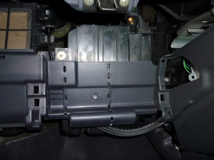 The image depicts a view inside the engine compartment of a vehicle, showing several parts of the car's internal system. The focus is on a silver metal component, which appears to be part of the dashboard or electronic controls. This section is partially disassembled, likely for repair or maintenance purposes. The presence of wires and connectors indicates that electrical connections are being examined or repaired. The overall setting suggests a professional repair or service environment.