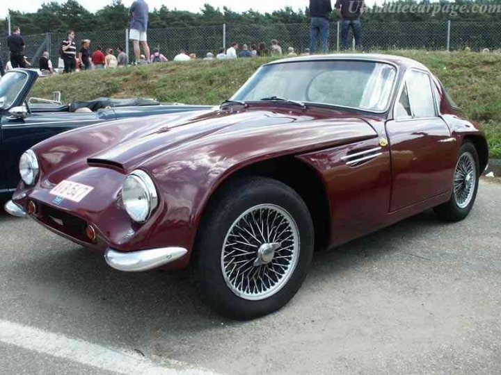 Early TVR Pictures - Page 35 - Classics - PistonHeads - The image displays a burgundy Aston Martin DB5 sports car parked on a paved road. The car's bright color and distinctive design features, such as the large headlights and chrome trimmings, are easily identifiable. There are a number of bystanders in the background, suggesting this may be a public event or gathering, perhaps a car show or a meet-up of people with similar vintage interests. The setting appears to be outdoors during the daytime.