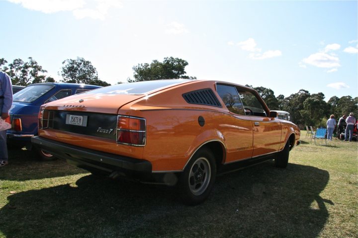 Pistonheads Americanaustralian - The image presents a vibrant scene in a bustling car show. The star of the show is an orange Datsun 280Z, its hue reminiscent of a retro volkswagen bug. The Datsun is parked on a lush grassy area, adjacent to a few other cars. The show seems to be held in a spacious outdoor location, with people milling about in the distance, their figures contrasting against the green of the surroundings. The blue sky overhead adds to the festive atmosphere, its brightness suggesting a perfect day for such an event.