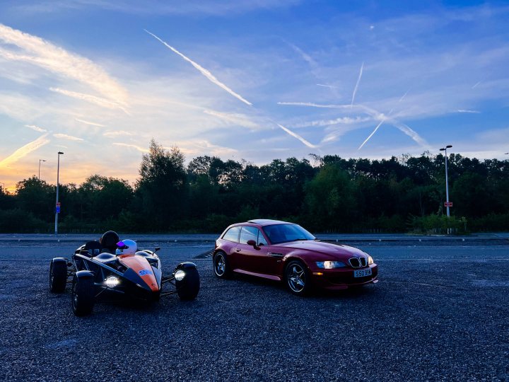 M Coupe Clownshoe, V8 M3 and 944 fun - Page 26 - Readers' Cars - PistonHeads UK - The image shows a gravel parking lot at twilight, with a clear sky indicating either dawn or dusk. In the foreground, there is a red sports car and two dirt bikes parked side by side, facing towards the right side of the image. On the far left, there appears to be a person standing near the sports car, although they are only partially visible. The setting suggests an outdoor event or gathering related to racing or motorsports. There is no visible text in the image.