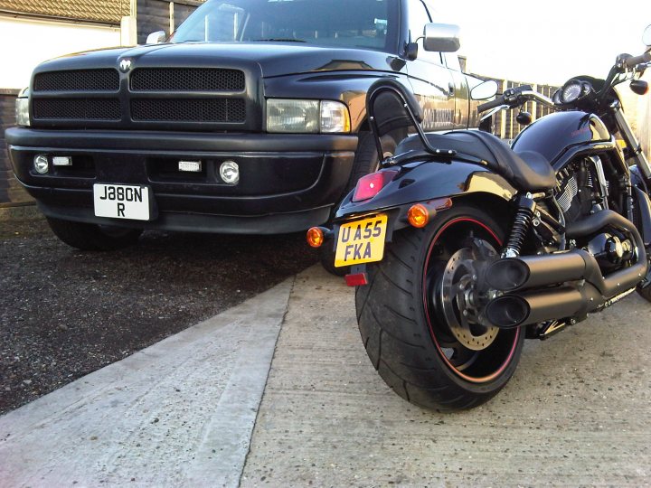 Pistonheads - The image shows a black motorcycle parked next to a black Dodge Ram pickup truck on a concrete lot. The motorcycle has a large rear wheel and is facing the viewer. The motorcycle displays the license plate "FKA" and has a yellow triangular headlight. The license plate on the Dodge Ram truck reads "J86 R."