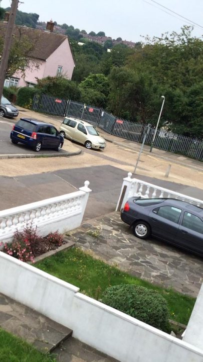 The BAD PARKING thread [vol3] - Page 42 - General Gassing - PistonHeads - The image presents a residential street scene with cars parked against a metal fence. There is a white car visible, and a driveway is also part of the scene, suggesting the presence of a home. The setting appears to be residential with a road curving in the background. The sky is overcast, suggesting a cloudy day. The grass in the foreground indicates that this is an outdoor residential area.