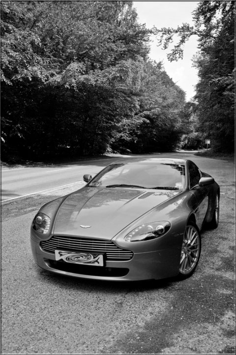 Pistonheads  Thread Aston - The image is a black and white photograph of a luxurious, long-nosed sports car with a sleek and aerodynamic design. It is parked on what appears to be a curved road or drive, with trees and foliage in the background, suggesting an environment away from densely populated urban areas. The car features a prominent grill and is seemingly well-maintained with a shiny exterior. The photo is somewhat grayscale and stylized, giving it a classic or vintage feel.