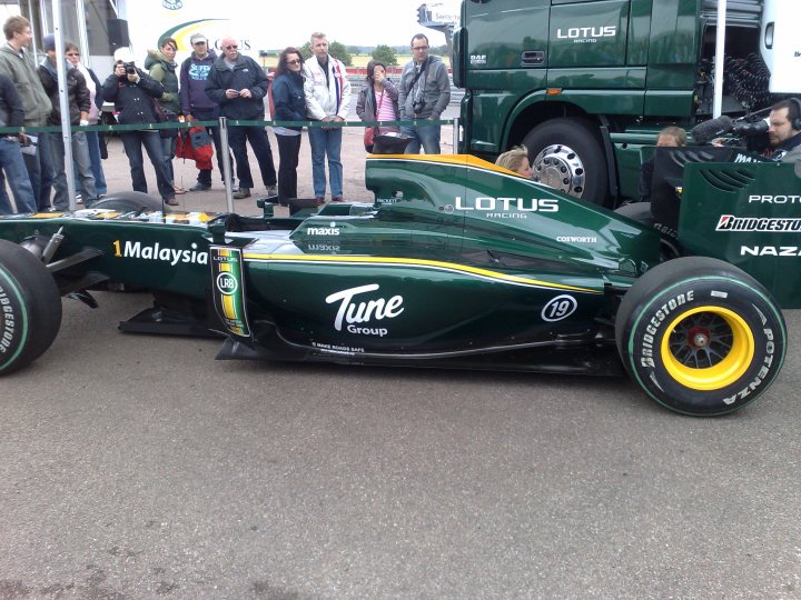 Pistonheads Snetterton - In the image, a vibrant green and yellow Lotus Formula 1 race car is parked outdoors, likely at an event or track. It's the central focus of the image, taking up a significant portion of the frame. The car has a sleek design and is adorned with various sponsor logos and details, including those for Tune, Proton, and Lotus.

Surrounding the car, a crowd of spectators is gathered, their attention captivated by the vehicle. Some of them are holding cameras and phones, possibly taking photos or recording the event. The scene suggests a public display or demonstration of the race car, likely at a trade show, auto show, or similar event.

The crowd varies in age, with individuals ranging from adults to children, all seemingly interested in the Formula 1 car on display. There's a palpable sense of excitement and interest in the air, a testament to the enduring appeal of motorsports.

One notable detail is the size and lighting of the Lotus logo on the car. These features stand out prominently against the car's green and yellow color scheme, making the Lotus branding a key element of the