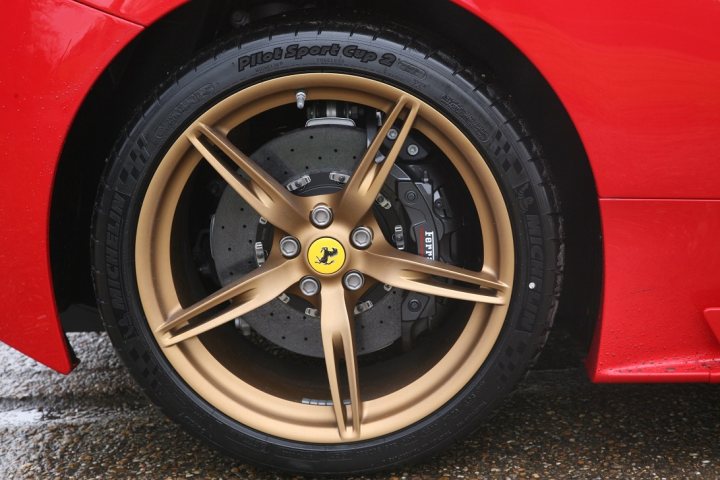 A close up of a black and silver motorcycle - Pistonheads - The image captures a close-up view of a black tire with bespoke gold-rimmed alloy wheels. The text on the tire indicates that it is a Ferrari tire. The wheels are designed with a classic star spokes pattern, adding a touch of luxury. The tire is set against a vibrant red background, possibly a sports car or Ferrari vehicle. The tire is horizontal with the wheel's center towards the top of the image, creating an interesting composition.