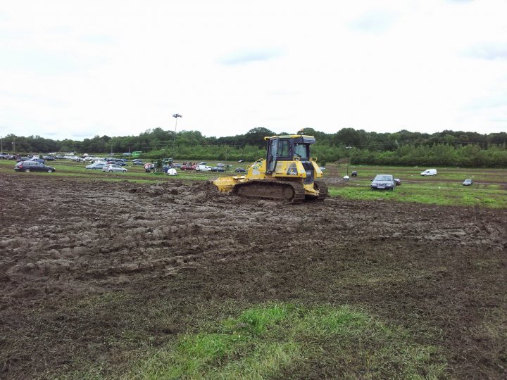 Silverstone F1 parking or park & ride? - Page 1 - Formula 1 - PistonHeads - This image depicts a large construction vehicle in the midst of mud, presumably engaged in excavation or groundwork. The vehicle is outdoors and appears to be in a muddy field or construction site. There are no visible texts or logos in the image. Hazy clouds are present in the top part of the image, indicating either early morning or late afternoon lighting conditions. There are several cars of various sizes in the background, some parked and others possibly on the move. The overall scene suggests a busy construction or industrial area. The image captures a sense of movement, with the construction vehicle clearly in focus.