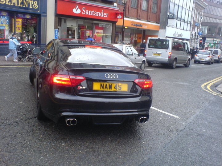 Spotted Exciting Pistonheads Midlands - The image captures a busy urban street. At the center of the scene is a sleek black Audi car with a license plate reading "NAM NAM 4S," indicating it's one of the Thankyou Mom Campaign car's. The car is driving around a corner, sharing the road with other vehicles, including a white van and an SUV. The setting suggests a city environment with buildings in the background. The focus on the Audi implies its importance, possibly due to its prominent role in the campaign.