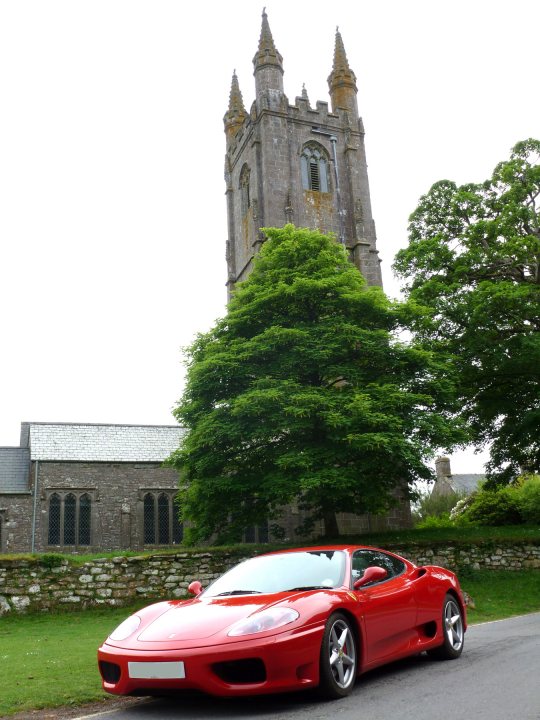 Isle of Arran in the Sunshine... - Page 1 - Supercar General - PistonHeads - The image shows a scene with a vibrant red sports car parked on a road, facing towards the right side of the image. The car is positioned in front of a building, which is an old church featuring a stone tower and multiple small windows. The tower has a detailed Gothic design and is topped with a pointed spire. There is a large, mature green tree growing close to the church and car, partially obscuring the view of the building. The sky appears overcast, creating a muted, soft-light ambiance to the scene. In the distance, there are more trees and the edge of additional buildings, suggesting a nearby residential area.