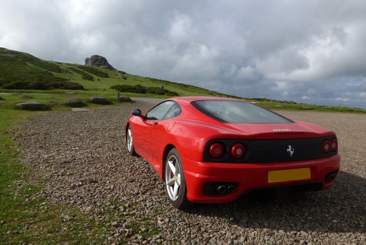 Isle of Arran in the Sunshine... - Page 1 - Supercar General - PistonHeads - The image captures a bright red convertible sports car positioned in the center of a rocky road. The car is sleek and modern, equipped with a large spoiler and a distinctive emblem on the back trunk. The backdrop of the image reveals a scenic view, with lush green hills dotted with trees and a meandering road that stretches into the distance. The combination of the vibrant red car, the natural terrain, and the clear sky creates a picturesque and serene scene.