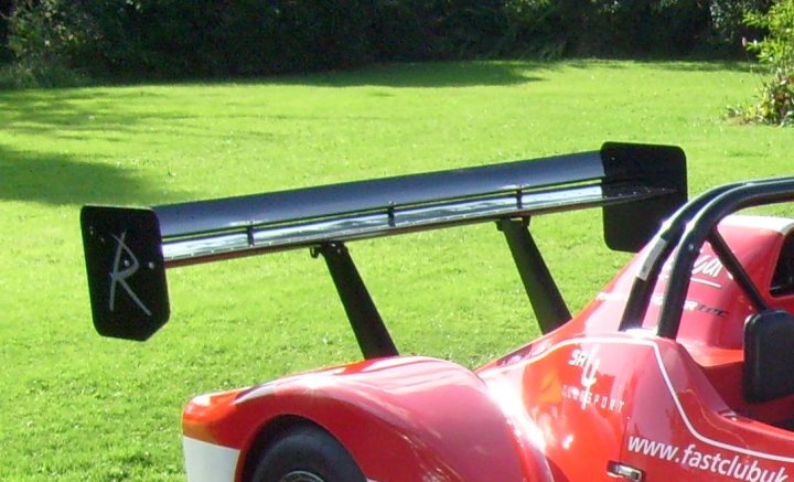 Biplane Pistonheads Wing - The image portrays a daring and sporty scene featuring a red, single-seat, racing go-kart. The vehicle's body is vibrant red, providing a stark contrast with the black windshield and equally black rear spoiler, both of which are significantly larger than usual, suggesting a design for aerodynamic performance. These elements are set against the backdrop of a serene, grassy field, hinting at a possible racing event or preparation for one. On the kart's side, the text "fastclubuk" is visible, possibly indicating a racing club or team affiliation.