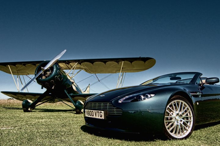 Pistonheads - The image features a black Aston Martin DDBRS car placed on a grassy field next to a vintage biplane. The car and plane are parallel to each other, creating a symmetrical composition. The sky is clear, allowing the viewer to see the vivid colors of the car and the plane's yellow wings. The placement of the car and plane suggests a juxtaposition of speed and history, with the car's sleek design contrasting against the biplane's historical and traditional aesthetic.