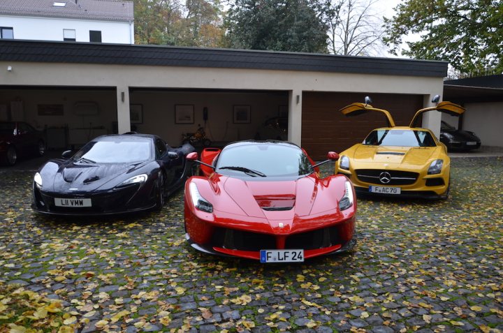 A red car is parked on the side of the road - Pistonheads - The image shows a trio of luxury sports cars, each parked on a brick driveway. The leftmost car is black, the middle one is red, and the rightmost one is yellow. They all feature sleek designs, long hoods, and short rear decks, characteristic of sports cars. The setting appears to be, at least partially, a residential area, given the presence of a house in the background, with a neatly arranged white-picket fence. The automotive craftsmanship and attention to detail indicate that these are high-end vehicles.