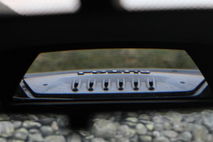 black evora spotted - Page 1 - Evora - PistonHeads - The image shows a close-up view of a rocky surface, likely the ground, through the open door of a vehicle, framed by a slightly cracked window. One can see the metal grille of the vehicle's ventilation system within the inset window, and outside of the vehicle, a glimpse of grass and the edge of a sunlit sky visible in the upper left corner, suggesting the vehicle is outdoors. The lower portion of the window is drooping slightly, creating a triangular composition through the glass.