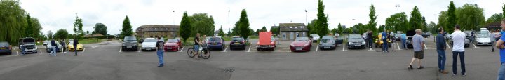 PH Meet - St Neots - Sunday 12th June - Page 2 - Herts, Beds, Bucks & Cambs - PistonHeads - The image is a panoramic shot taken outside, possibly in a parking lot. There are multiple lines of parked cars, showcasing an array of colors and types. In the foreground, there are visible motion blur effects on two figures, possibly due to the camera's movement at the time the photo was taken. The spot where the cars are parked appears to be full and somewhat compact. The image captures a typical urban scene, filled with the everyday hustle and bustle of vehicles and people.