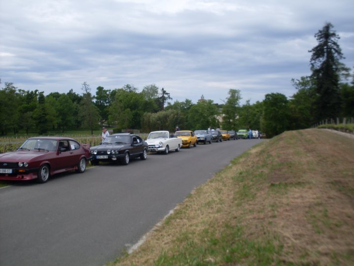 Vacance de France refroidis par air... - Page 2 - Classic Cars and Yesterday's Heroes - PistonHeads - The image captures a line of vintage cars parked along the side of a road. The cars are diverse in color, dominated by yellow, white, black, and red. They are arranged in a line that creates a pleasing procession of shapes and colors. The perspective of the photo is from the top, giving a bird's-eye view of the road and the cars, as well as some visible greenery in the background. The sky peeks from the top left of the image, softening the scene with its light color.