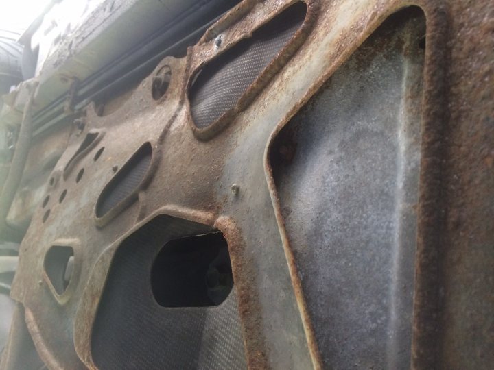 Help Needed! Mk 2.5 Rust - Page 1 - Mazda MX5/Eunos/Miata - PistonHeads - The image presents a close-up view of an engine cover, likely made of metal or a metal alloy. The cover is filthy and rusted, suggesting it has been exposed to the elements for a long time. The engine cover has several holes in it, one of which is quite large, seemingly to house a probe or some other mechanical component. The backdrop is blurry, with hints of machinery or mechanical components that are of a different color, possibly indicating they are made of aluminum or another light-colored metal. The overall image exudes a sense of rustic industrial abandonment.