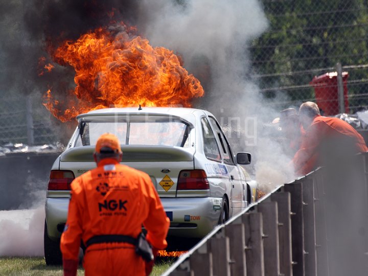 Recommend me a Fire Extinguisher please :) - Page 1 - UK Club Motorsport - PistonHeads - The image depicts a dramatic scene of a car engulfed in flames. The car is parked near a safety barrier, suggesting it might be at a racetrack. There are two people visible in the image, both seen from the back. One is likely the driver, while the other could be a member of the safety team or a bystander. The surrounding area is filled with smoke, and the fire appears to be intense, with flames shooting high and brightly against the sky. The overall image conveys a sense of urgency and potential danger.