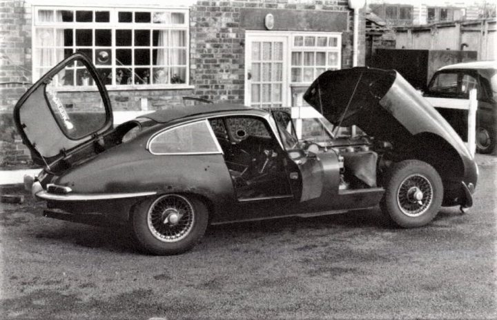 How about a 'period' classics pictures thread - Page 475 - Classic Cars and Yesterday's Heroes - PistonHeads - The image shows a classic sports car with its hood open, next to an old-fashioned brick building with a contrasting white door and window frames. The car has a distinctive design with long wheel arches, a short front end, and iconic round taillights. An umbrella is open, seemingly held by an unseen person, beside the car, suggesting rain or preparation for it. The black and white format of the photo gives it a timeless, vintage feel.