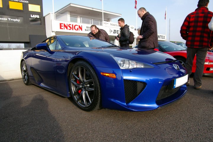 Goodwood Breakfast Club - Sushi Sunday - photos - Page 1 - Goodwood Events - PistonHeads - The image shows a bright blue hybrid sports car parked in what appears to be a showroom or dealership setting. Four men are standing around the car, examining it with interest. The car has a sleek design with sporty proportions and is positioned so that the side profile is clearly visible. The dealership or showroom exhibits a modern and professional atmosphere with signage displaying brand names like Ensign and Lexus.