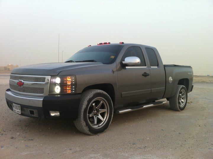 Not what  I had in mind but ....... - Page 1 - Yank Motors - PistonHeads - The image captures a scene of a desolate landscape. A gray Chevrolet truck, with its large grille and chrome accents, is parked on a dirt road. The truck's four red tail lights are prominently displayed against the dusky sky. In the background, a large metal structure with a red glare can be seen. The sky above the truck is a hazy orange, suggesting either a sunrise or sunset.