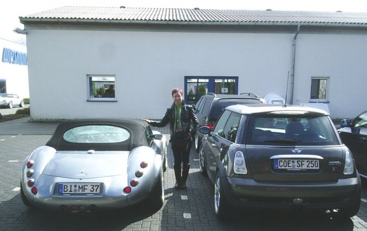 Classics dwarfed by moderns - Page 32 - Classic Cars and Yesterday's Heroes - PistonHeads - This image captures a woman standing next to two unique cars in a parking lot. The first car has a cylindrical body and is made of metal, while the second car is a small black SUV. The woman is wearing a green scarf and is smiling at the viewer. In the background, there is a modern building with a blue door. The overall scene suggests a relaxed and sunny day.