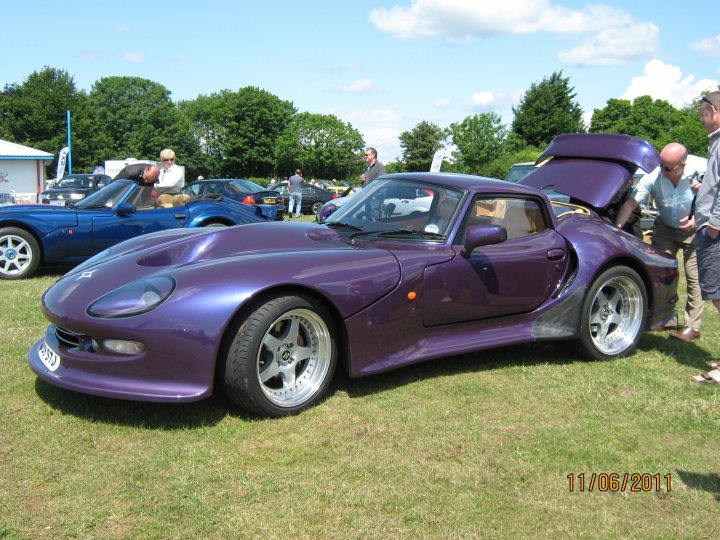 Pistonheads - This image showcases a trio of vibrant sports cars displayed on a grassy field under a partly cloudy sky. The most prominent is a sleek purple sports car parked centrally, adorned with silver wheels and a prominent front grille. Slightly behind and to the left, there's a blue sports car, also featuring silver wheels, adding a contrast to the purple car. In the background, behind a collection of other cars, two individuals can be seen, possibly admiring the display. The vehicles are away from buildings, suggesting an outdoor car show or similar event.