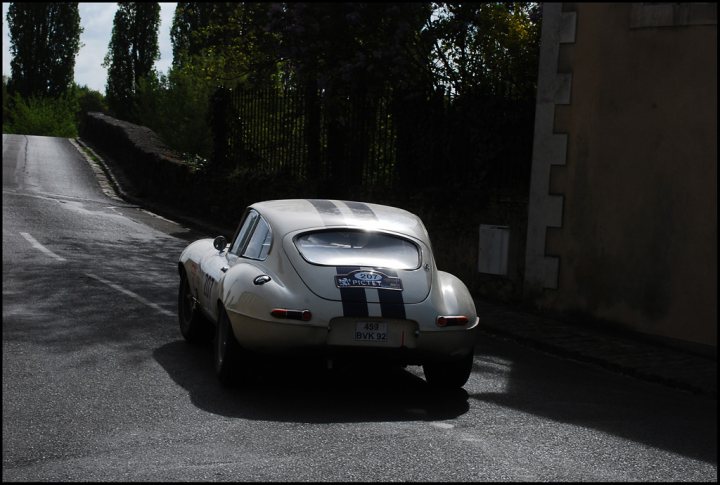 The Tour Auto came to town - Page 1 - Classic Cars and Yesterday's Heroes - PistonHeads - The image shows a vintage-style race car driving down a narrow rural road. The car has a classic design with a long body, a prominent rear spoiler, and a striped hood which are common features of touring and racing cars from the past. The car is casting a shadow behind it, indicating that the sun is shining from the left side of the photo and the weather seems to be clear. The road itself is bordered by a stone wall on the right side and vegetation on the left, adding to the countryside ambiance. The setting suggests a peaceful, rural location in contrast to the high-performance nature of the car.