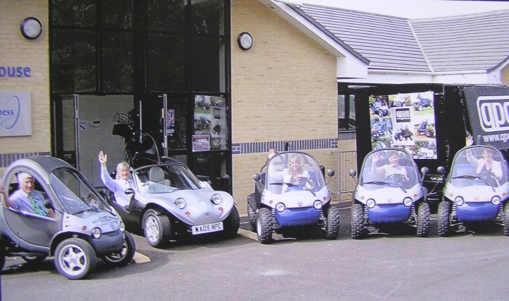 A group of motorcycles parked in front of a building - Pistonheads - The image features a group of people sitting in small, cart-like vehicles parked outside a building. There are three such vehicles visible, and the individuals appear to be enjoying their time inside these quirky rides. The setting suggests a special event or exhibition, as evidenced by the sign on the building that reads "house". The atmosphere seems lively, with one person in particular waving towards the camera. The presence of a truck and another individual indicates the public nature of this gathering.