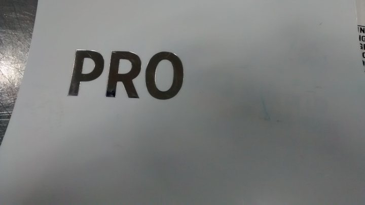 Everywhere I look........... - Page 4 - Le Mans - PistonHeads - The image shows a close-up of a white surface with the word "PRO" prominently in the center. The "PRO" is written in large, bold letters with a metallic foil finish, giving it a slightly three-dimensional effect. The background appears blurry, emphasizing the text. The font is sans-serif, which gives it a modern and clean appearance. The lighting on the text seems to reflect light, suggesting it could be on a glossy surface.