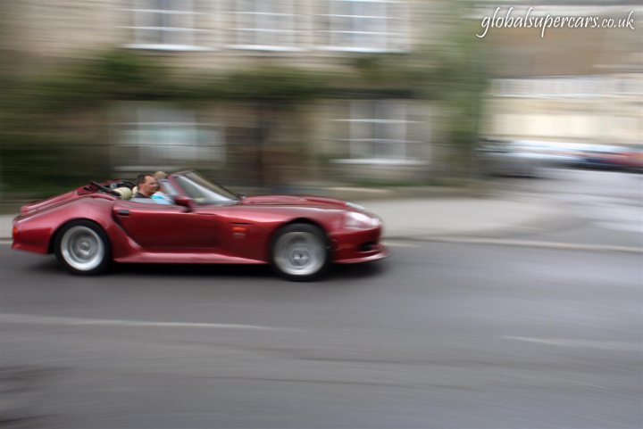 Sunday April 25th Bradford on Avon - Marocs - Page 1 - South West - PistonHeads - The image shows a red sports car speeding down a wet street, as indicated by the glistening road surface. The vehicle is captured in motion, with its dynamic force blurring the wheel on the driver's side. In the background, there are apartment buildings, suggesting an urban residential setting. In the lower corner of the image, there's a watermark with the text "Global Supersports.co.uk," indicating the source or website associated with the image.