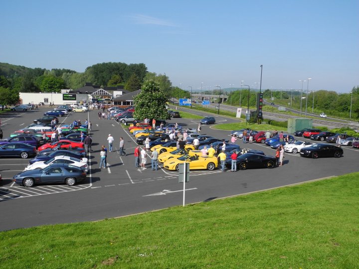 Gordano this morning en route to cwc - Page 1 - South West - PistonHeads - The image displays a vibrant scene at a parking lot, bathed in the warm hues of the daytime sun. A variety of vehicles, including cars and a truck, are parked in several rows. The vehicles showcase a range of colors and models, adding dynamism to the otherwise static scene. Background elements like trees and a building hint at a suburban or semi-urban setting. A group of people can be seen strolling around, lending a sense of scale and activity to the image. The perspective from an elevated angle provides a comprehensive view of the parking lot, showcasing its layout and the multitude of objects within.