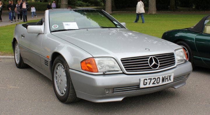 500sl r129 - First 6 months - Page 1 - Mercedes - PistonHeads - The image captures a quaint outdoor scene with a focus on a silver Mercedes convertible parked on the side of a road. The car has a European license plate on the front, and is an older model, with the top down, revealing the classic design. In the background, there are people strolling along the sidewalk and other cars parked. The presence of the license plate and the car's style suggest that this might be a European city or town.