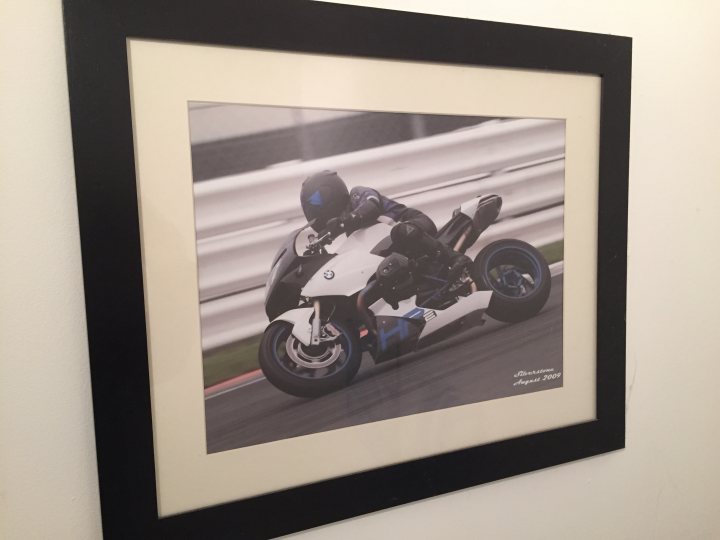 Art on your walls... - Page 14 - Homes, Gardens and DIY - PistonHeads - The image shows a framed photograph of a person riding a motorcycle on a race track. The rider is wearing a full racing attire, including a helmet and leather suit, and is tilted to one side suggesting rapid movement. The motorcycle is in motion on a track, visible in the background, with green and white grass and white guardrails along its length. The photograph is displayed on a wall with a black frame giving it a modern art vibe. A person's ankle is visible in the bottom right corner of the photograph, indicating that it might have been taken from a lower viewpoint or during a celebration of the race.