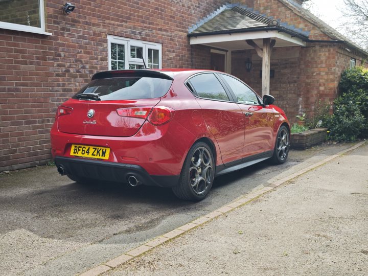 Alfa Romeo Giulietta Quadrifoglio Verde (Launch edition)  - Page 4 - Readers' Cars - PistonHeads UK - The image features a red car parked on the side of a driveway. It has a sporty look with a black roof rack and a black exhaust pipe. There are two black wheel rims, suggesting it's well-maintained or possibly customized for performance. The license plate is clearly visible, adding to the realism of the image. The car is parked in front of a house, which has a white facade and a brick chimney, suggesting a residential area.