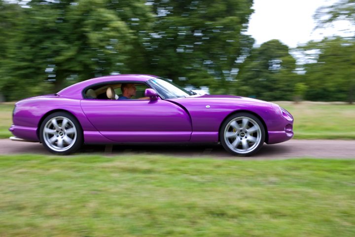 Motors Pistonheads Manor - In the image, we see a vibrant purple Spyker, a small two-seater luxury sports car, cruising down a road. Two individuals are inside the car, enjoying the ride. The car is giving off a sense of speed, which is further enhanced by the blurred effect on the grasses and trees lining the road. The car and its occupants are the main focus of the image, while the road and surroundings provide a blue sky backdrop. This image encapsulates a dynamic moment of speed and luxury, set against the serene beauty of nature.