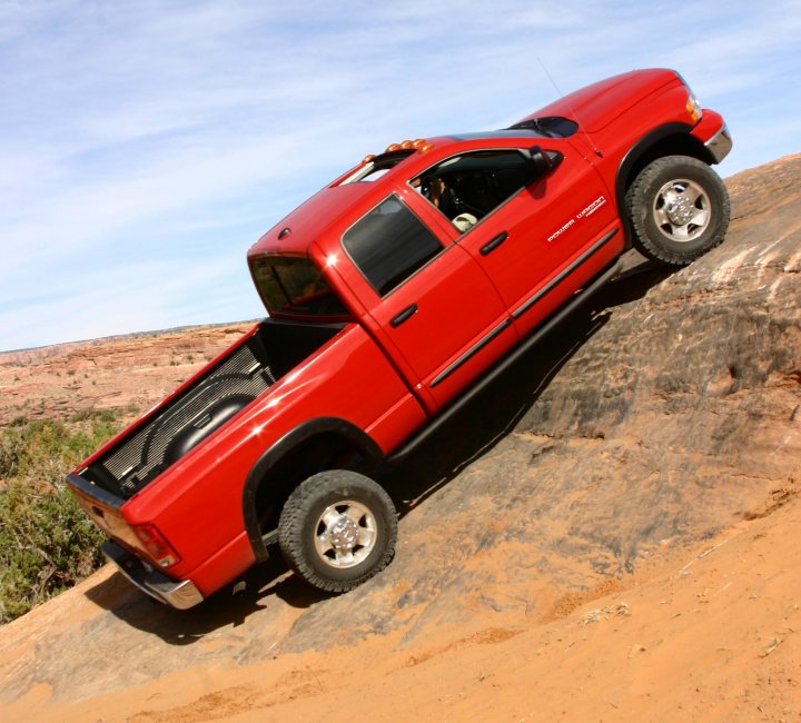 Anyone Driven A Dodge Ram Off Road? - Page 1 - Off Road - PistonHeads - The image shows a red Dodge Ram pickup truck that appears to be stuck on a rocky outcropping. The truck is viewed from a low angle directly underneath it, emphasizing the height of the elevation it's on. The setting suggests a desert or similarly arid environment, with dry earth and sparse vegetation. The truck is equipped with a toolbox in the bed and all-terrain tires, indicating it's prepared for off-road conditions.