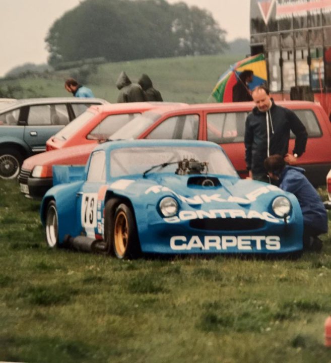 Early TVR Pictures - Page 114 - Classics - PistonHeads - This image captures a fascinating scene at an outdoor event. Four cars are parked in a grassy lot, with their open hoods revealing detailed engines. The colors of the cars are vibrant: blue, red, and black. The atmosphere is one of curiosity, with blankets spread out on the ground and several people milling about, inspecting the cars. The setting suggests a gathering of car enthusiasts.