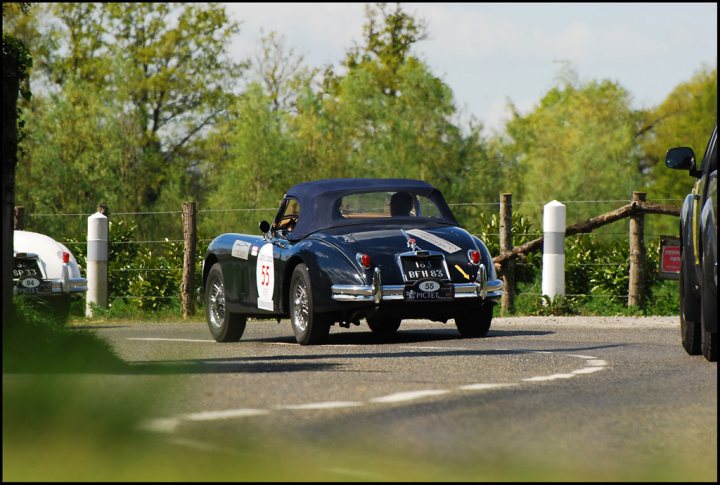 The Tour Auto came to town - Page 1 - Classic Cars and Yesterday's Heroes - PistonHeads - In the image, a vintage blue car with a white top is driving down an open road. The road stretches out ahead of the car, lined with a wooden fence and thick trees on both sides. The sun is shining brightly, casting long shadows that add to the picturesque scene. The background features an additional vehicle from the 1940s era, suggesting a classic vehicle event or exhibit.